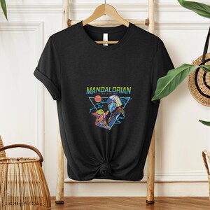 May include: Black t-shirt with a vibrant graphic of The Mandalorian and Grogu. The design includes the text "MANDALORIAN" and "THIS IS THE WAY" within a triangle, set against a sunset and space backdrop. The t-shirt is displayed on a wooden hanger.
