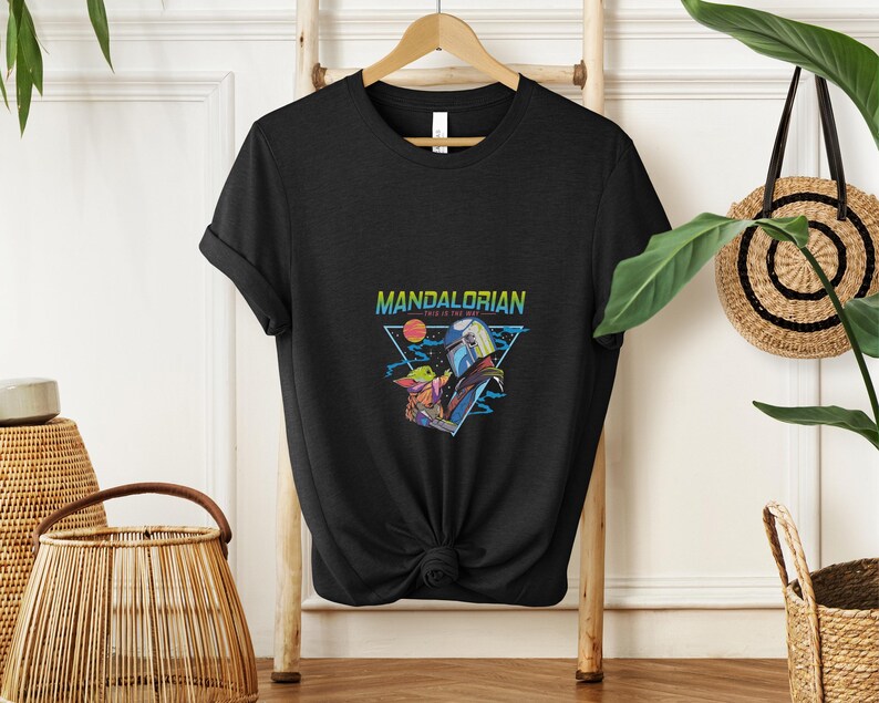 May include: Black t-shirt with a vibrant graphic of The Mandalorian and Grogu. The design includes the text "MANDALORIAN" and "THIS IS THE WAY" within a triangle, set against a sunset and space backdrop. The t-shirt is displayed on a wooden hanger.