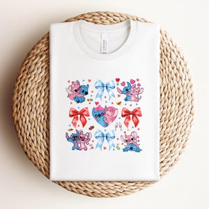 May include: White t-shirt featuring a colorful graphic of cartoon characters, hearts, and bows. The design includes pink and blue characters in various poses, red and blue bows, and heart accents. The shirt is folded on a woven surface.