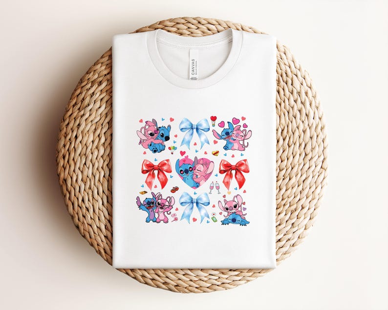 May include: White t-shirt featuring a colorful graphic of cartoon characters, hearts, and bows. The design includes pink and blue characters in various poses, red and blue bows, and heart accents. The shirt is folded on a woven surface.