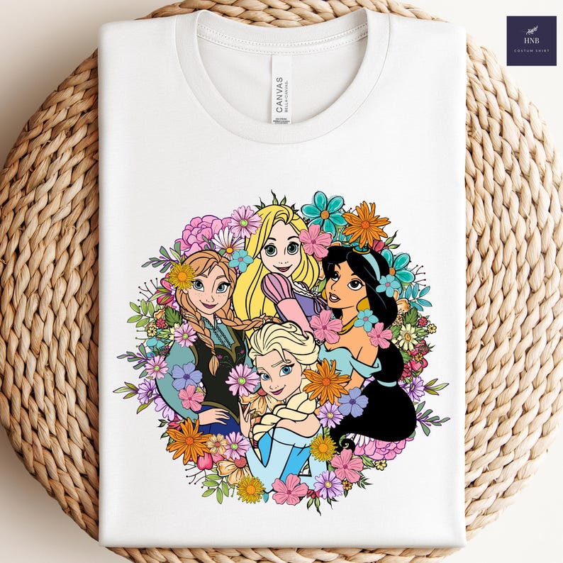 May include: White t-shirt featuring a colorful graphic of animated princesses surrounded by flowers. The design includes characters with blonde, black, and auburn hair, set against a floral backdrop. The shirt has a crew neck and short sleeves.