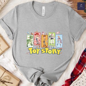 Puede incluir: Una camiseta gris jaspeada con las palabras "Toy Story" en amarillo y azul. La camiseta muestra im&aacute;genes rectangulares coloridas de personajes de la pel&iacute;cula animada, incluyendo un dinosaurio verde, Woody, Buzz Lightyear, y otros. La camiseta es de material suave.