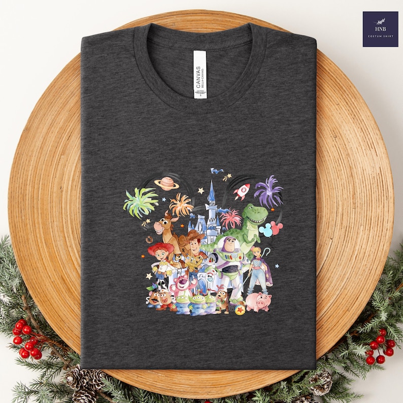 May include: Dark gray t-shirt featuring a colorful illustration of characters from the animated film "Toy Story." The design includes Woody, Buzz Lightyear, and other characters, with fireworks and a castle in the background. The shirt is folded on a wooden tray.