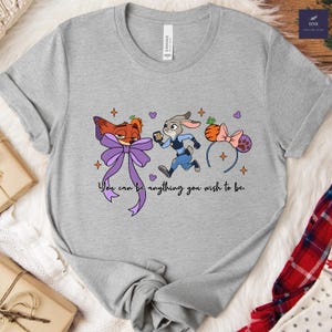 May include: A gray t-shirt featuring cartoon characters from Zootopia. The design includes Nick Wilde with a purple bow, Judy Hopps running, and a carrot headband. The text reads, "You can be anything you wish to be."