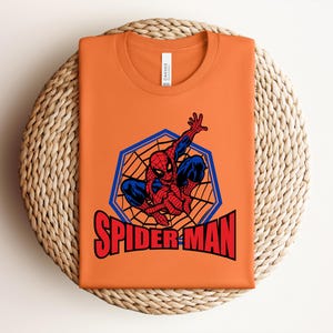 May include: Orange t-shirt featuring a Spider-Man graphic. The design includes Spider-Man in a web, with the words "SPIDER-MAN" in red. The shirt is folded on a woven, circular surface.