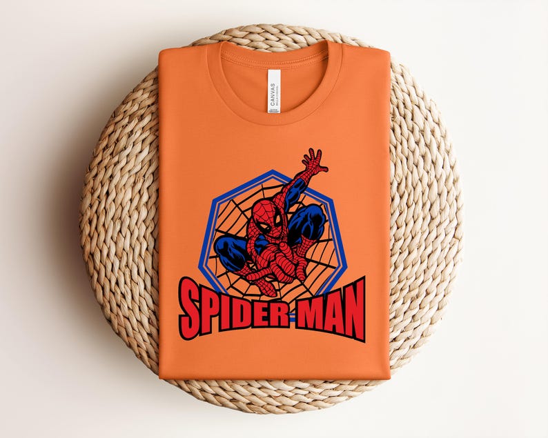May include: Orange t-shirt featuring a Spider-Man graphic. The design includes Spider-Man in a web, with the words "SPIDER-MAN" in red. The shirt is folded on a woven, circular surface.