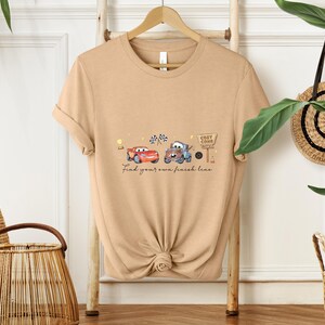 May include: Tan t-shirt featuring a watercolor design of two cartoon cars, a red one and a blue one, with checkered flags and a sign that says "Cozy Cone Motel." The text "Find your own finish line" is below the cars.