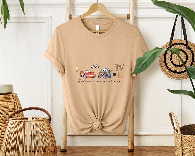 May include: Tan t-shirt featuring a watercolor design of two cartoon cars, a red one and a blue one, with checkered flags and a sign that says "Cozy Cone Motel." The text "Find your own finish line" is below the cars.