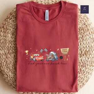 May include: A maroon t-shirt featuring a graphic of two cartoon cars, a trophy, and checkered flags. The text "Find your own finish line" is written below the cars. The shirt has a round neckline.