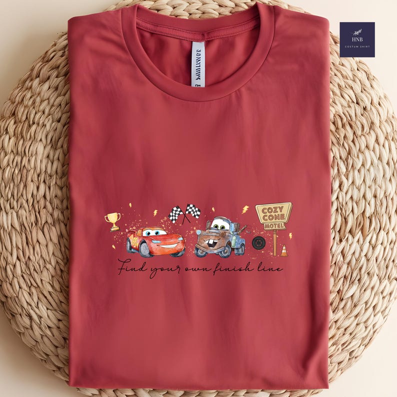 May include: A maroon t-shirt featuring a graphic of two cartoon cars, a trophy, and checkered flags. The text "Find your own finish line" is written below the cars. The shirt has a round neckline.