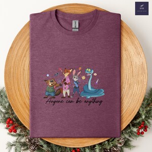 K&ouml;nnte beinhalten: Burgunderfarbenes T-Shirt mit Cartoon-Figuren und dem Text "Anyone can be anything". Die Grafik zeigt einen Biber, einen Fuchs, ein Kaninchen und eine Schlange. Das Shirt liegt auf einem Holztablett mit Weihnachtsdekoration.