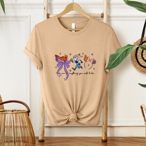 May include: Tan t-shirt featuring a cartoon design with a fox, a rabbit, and mouse ears. The text "You can be anything you wish to be" is written in a script font. The shirt is hanging on a wooden hanger.