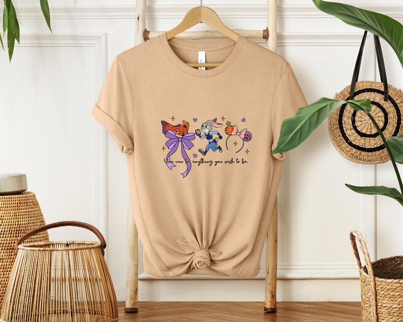 May include: Tan t-shirt featuring a cartoon design with a fox, a rabbit, and mouse ears. The text "You can be anything you wish to be" is written in a script font. The shirt is hanging on a wooden hanger.
