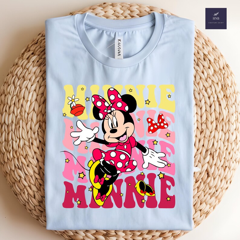 May include: Light blue t-shirt featuring a colorful graphic of Minnie Mouse. The design includes Minnie in a red polka dot dress and bow, with the name "Minnie" in layered retro fonts. The shirt is folded on a woven surface.