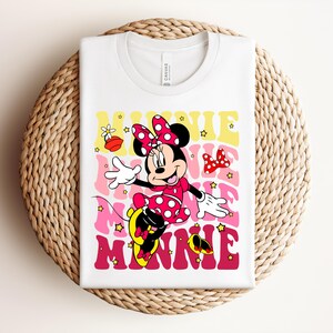 May include: White t-shirt featuring a retro design of Minnie Mouse in a pink polka-dot dress and bow. The word "MINNIE" is printed in a layered, colorful font. The design includes stars and a bow.