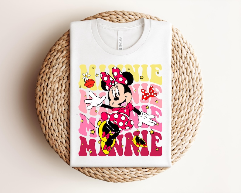 May include: White t-shirt featuring a retro design of Minnie Mouse in a pink polka-dot dress and bow. The word "MINNIE" is printed in a layered, colorful font. The design includes stars and a bow.