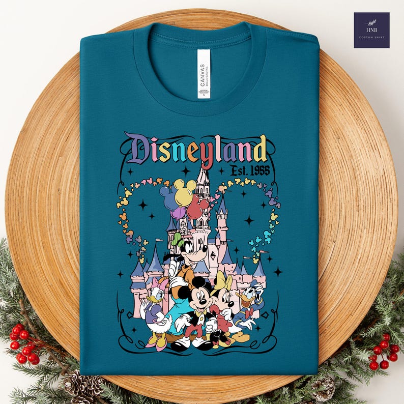 Może przedstawiać: Turkusowy t-shirt z okrągłym dekoltem i grafiką zamku Disneyland z Myszką Miki, Myszką Minnie, Kaczorem Donaldem, Daisy i Goofym. Napis brzmi Disneyland Est. 1955, otoczony kolorowymi balonami w kształcie głowy Miki oraz gwiazdkami.