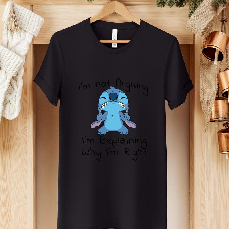 May include: Black t-shirt featuring a cartoon character with the text "I'm not Arguing, I'm Explaining Why I'm Right". The character is blue with large ears and a grumpy expression. The shirt is hanging on a wooden hanger.