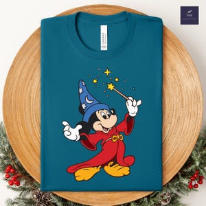 Puede incluir: Camiseta de color turquesa con un dise&ntilde;o de Mickey Mouse de dibujos animados como mago. Mickey lleva un sombrero azul y una t&uacute;nica roja, sosteniendo una varita con estrellas. La camiseta tiene una etiqueta "Canvas".