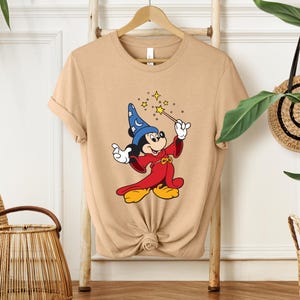 Puede incluir: Camiseta color beige con Mickey Mouse de dibujos animados vestido de mago. Mickey lleva un sombrero de mago azul con estrellas blancas, una t&uacute;nica roja y zapatos amarillos. Sostiene una varita con estrellas brillantes.