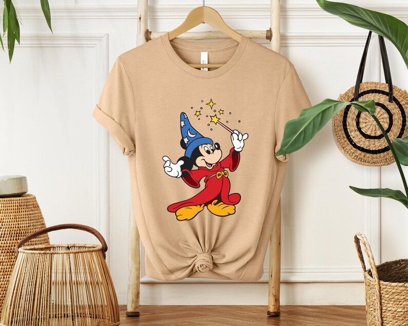 Puede incluir: Camiseta color beige con Mickey Mouse de dibujos animados vestido de mago. Mickey lleva un sombrero de mago azul con estrellas blancas, una t&uacute;nica roja y zapatos amarillos. Sostiene una varita con estrellas brillantes.