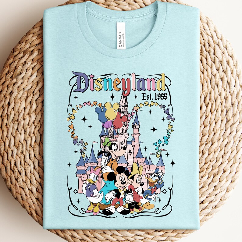 Może przedstawiać: Jasnoniebieski t-shirt z nadrukiem graficznym Disneylandu i napisem "Disneyland Est. 1955". Wz&oacute;r przedstawia zamek, Myszkę Miki, Myszkę Minnie, Goofy'ego, Kaczora Donalda i Daisy, otoczonych kolorowymi balonami.