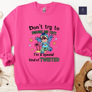 May include: A vibrant pink sweatshirt featuring a graphic of a blue cartoon character with the text "Don't try to FIGURE ME OUT, I'm a special kind of TWISTED." The design includes palm trees and stars.