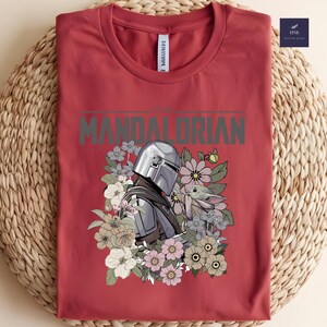 Puede incluir: Camiseta roja con un dise&ntilde;o de The Mandalorian y Grogu rodeados de flores. El texto "THE MANDALORIAN" est&aacute; impreso en gris. La camiseta est&aacute; doblada sobre una superficie tejida.