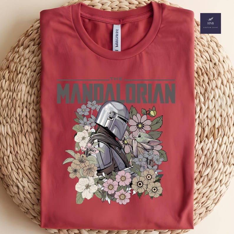 Puede incluir: Camiseta roja con un dise&ntilde;o de The Mandalorian y Grogu rodeados de flores. El texto "THE MANDALORIAN" est&aacute; impreso en gris. La camiseta est&aacute; doblada sobre una superficie tejida.