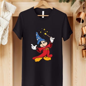 Puede incluir: Camiseta negra con una ilustraci&oacute;n de dibujos animados de Mickey Mouse como hechicero. Mickey lleva un sombrero azul, una t&uacute;nica roja y zapatos amarillos, sosteniendo una varita con estrellas. El dise&ntilde;o est&aacute; centrado en la camiseta.