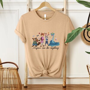 K&ouml;nnte beinhalten: Beigefarbenes T-Shirt mit einer Cartoon-Illustration von Tierfiguren: einem Biber, einem Fuchs, einem Kaninchen und einer Schlange. Der Text "Anyone can be anything" steht darunter.