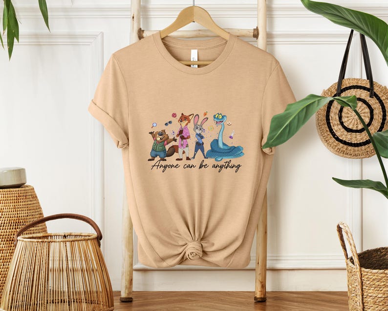 K&ouml;nnte beinhalten: Beigefarbenes T-Shirt mit einer Cartoon-Illustration von Tierfiguren: einem Biber, einem Fuchs, einem Kaninchen und einer Schlange. Der Text "Anyone can be anything" steht darunter.