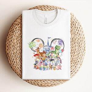 May include: White t-shirt featuring a colorful illustration of various characters. The design includes a castle, palm trees, and the word "Toy". The shirt is made of a soft material and is displayed on a woven surface.