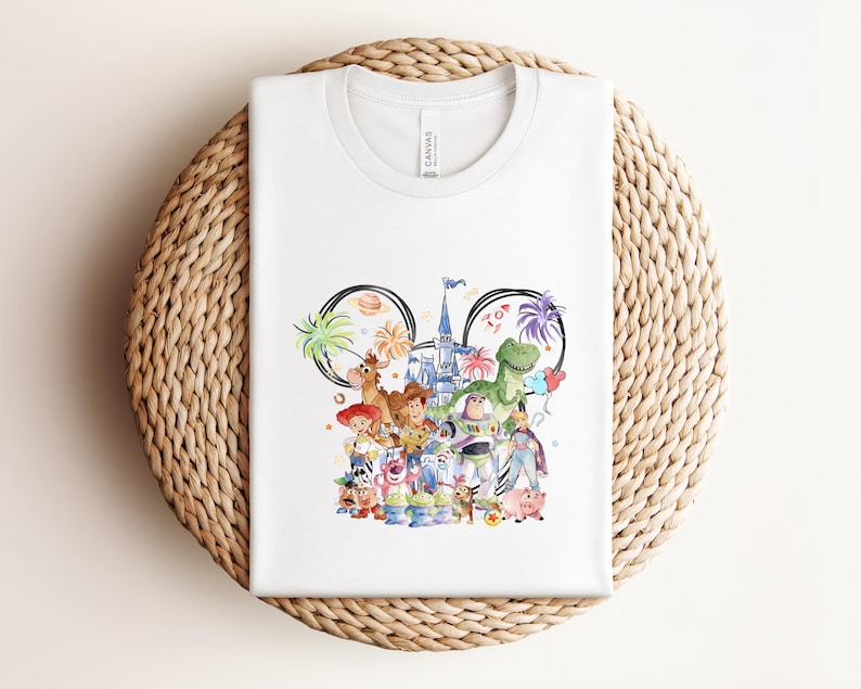May include: White t-shirt featuring a colorful illustration of various characters. The design includes a castle, palm trees, and the word "Toy". The shirt is made of a soft material and is displayed on a woven surface.