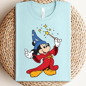 Puede incluir: Camiseta azul claro con una ilustraci&oacute;n de dibujos animados de Mickey Mouse como mago. Mickey lleva un sombrero de mago azul con estrellas blancas, una t&uacute;nica roja y zapatos amarillos. Sostiene una varita con una estrella, rodeada de otras estrellas.