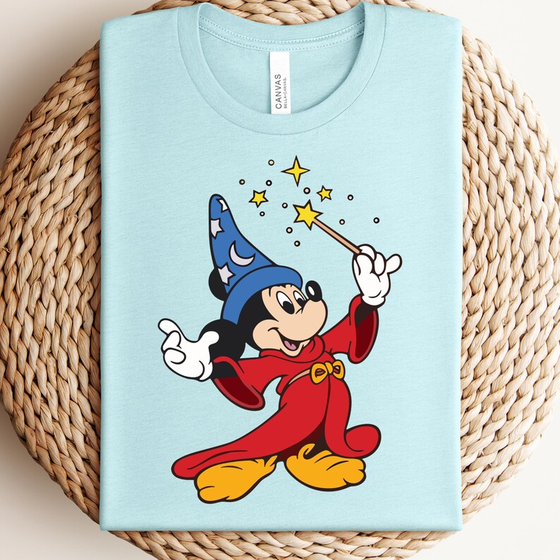 Puede incluir: Camiseta azul claro con una ilustraci&oacute;n de dibujos animados de Mickey Mouse como mago. Mickey lleva un sombrero de mago azul con estrellas blancas, una t&uacute;nica roja y zapatos amarillos. Sostiene una varita con una estrella, rodeada de otras estrellas.