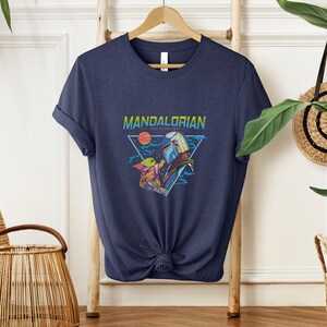 May include: Navy blue t-shirt with a graphic from the Mandalorian series. The design includes the text "MANDALORIAN" and "THIS IS THE WAY" with illustrations of characters and a space scene. The shirt is hanging on a wooden hanger.
