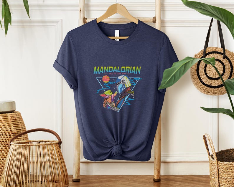 May include: Navy blue t-shirt with a graphic from the Mandalorian series. The design includes the text "MANDALORIAN" and "THIS IS THE WAY" with illustrations of characters and a space scene. The shirt is hanging on a wooden hanger.