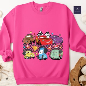 May include: A bright pink sweatshirt featuring a colorful graphic of cartoon cars. The design includes various car characters, checkered flags, stars, and a trophy. The sweatshirt is made of a soft material.