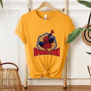 May include: Mustard yellow t-shirt featuring a Spider-Man graphic. The graphic shows Spider-Man in a web with the words "SPIDER-MAN" in red letters. The shirt is hanging on a wooden hanger.