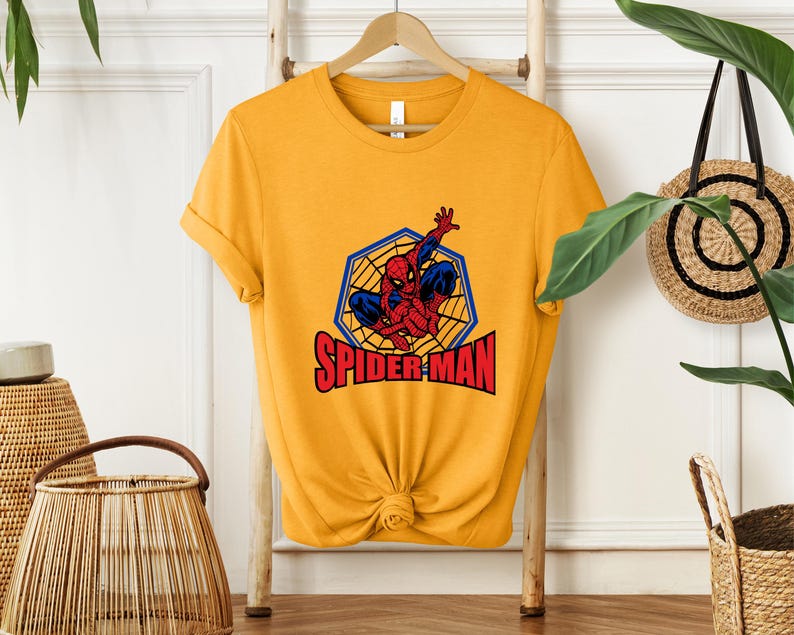 May include: Mustard yellow t-shirt featuring a Spider-Man graphic. The graphic shows Spider-Man in a web with the words "SPIDER-MAN" in red letters. The shirt is hanging on a wooden hanger.