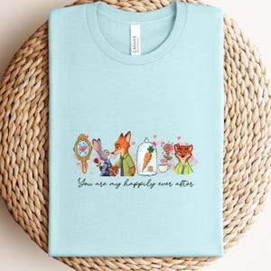 May include: A light blue t-shirt featuring a cartoon design with characters from Zootopia. The design includes a mirror, bunny, fox, carrot, and roses. The text "You are my happily ever after" is written below the design.