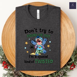 May include: Dark gray t-shirt featuring a cartoon character with the text "Don't try to figure me out, I'm a special kind of twisted." The character is blue with a floral lei and tropical shorts. The shirt is folded.
