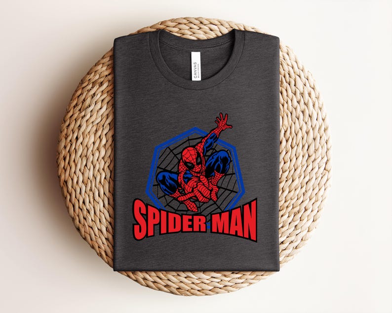 May include: Dark gray t-shirt featuring a Spider-Man graphic. The design includes a red and blue Spider-Man figure within a blue web-like frame, with the words "SPIDER MAN" in red block letters below. The shirt is folded on a woven surface.