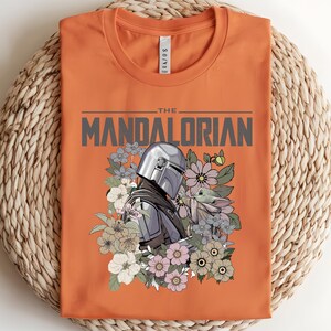 Puede incluir: Camiseta naranja con un gr&aacute;fico de The Mandalorian y Grogu rodeados de flores. El texto "THE MANDALORIAN" est&aacute; impreso en la parte superior. La camiseta est&aacute; doblada sobre una superficie tejida.