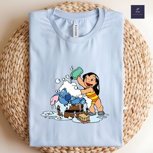 Pode incluir: Camiseta azul clara com uma ilustra&ccedil;&atilde;o de desenho animado de Lilo e Stitch. Lilo est&aacute; jogando &aacute;gua em Stitch, que est&aacute; em uma banheira de madeira. A camiseta tem a palavra "ENCOVER" impressa.