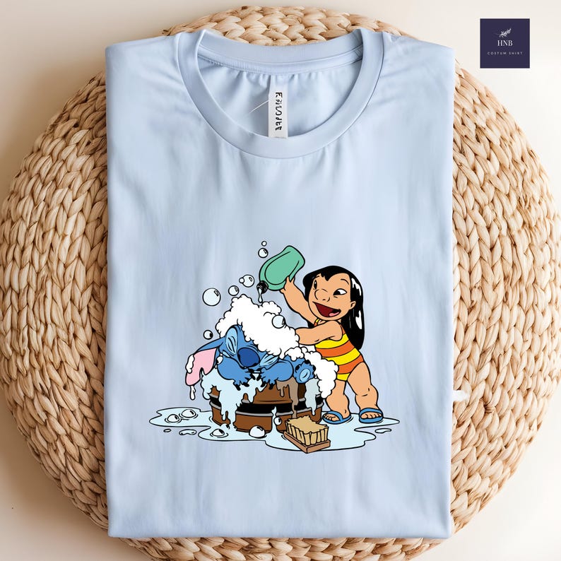 Pode incluir: Camiseta azul clara com uma ilustra&ccedil;&atilde;o de desenho animado de Lilo e Stitch. Lilo est&aacute; jogando &aacute;gua em Stitch, que est&aacute; em uma banheira de madeira. A camiseta tem a palavra "ENCOVER" impressa.