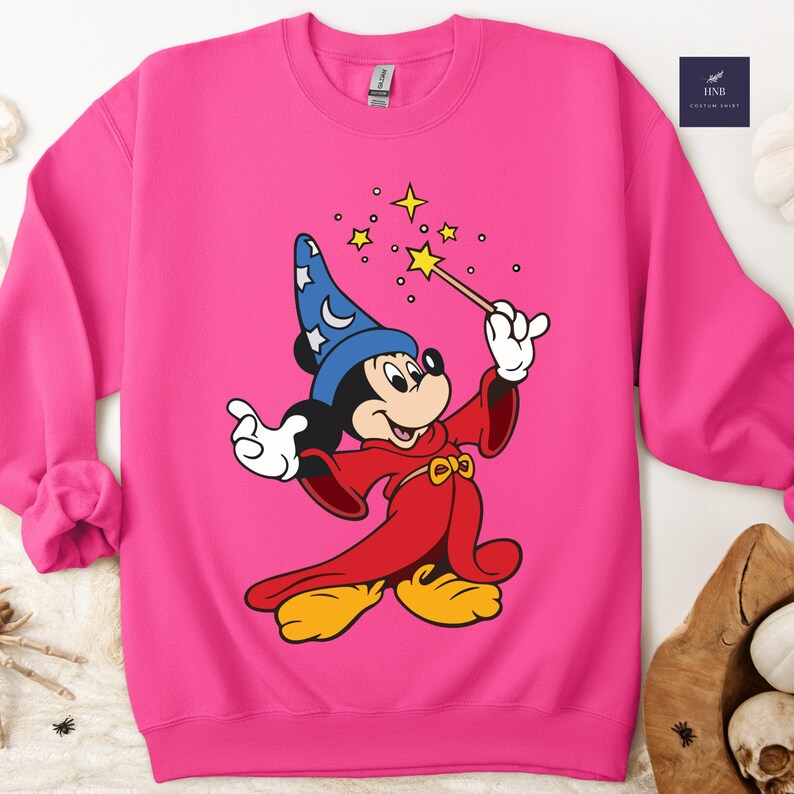Puede incluir: Sudadera rosa brillante con una ilustraci&oacute;n de dibujos animados de Mickey Mouse vestido de mago. Mickey lleva un sombrero de mago azul con estrellas, una t&uacute;nica roja y una varita con una estrella. El dise&ntilde;o incluye estrellas amarillas.