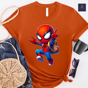 Puede incluir: Camiseta naranja con un personaje de dibujos animados de Spider-Man haciendo un gesto de rock. El superh&eacute;roe lleva un traje rojo y azul y tiene una mochila. La camiseta tiene cuello redondo y mangas cortas.
