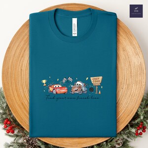 May include: Teal t-shirt featuring a graphic of two cartoon cars, a trophy, and the words "Find your own finish line." The design includes a Cozy Cone Motel sign and checkered flags.